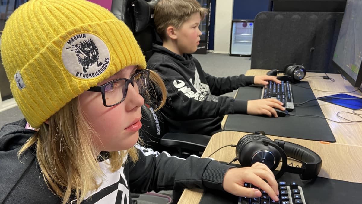 Kaksi alle pikkupoikaa istuu vierekkäin tietokoneiden äärellä pelaamassa tietokonepelejä. Etummaisella on keltainen pipo päässä ja silmälasit