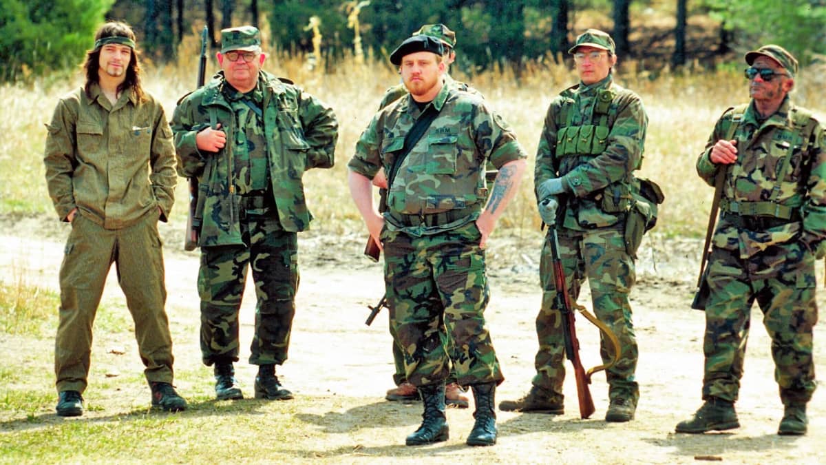 Joukko miehiä erilaisissa vihreissä maastoasuissa seisoo aukiolla aseiden kanssa.