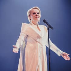 Sinivalkoinen ääni Katri Helena UMK-lavalla mikrofonin edessä. Helenalla on yllään valkoinen mekko ja tausta on tumma.