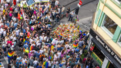 Suuri joukko ihmisiä kadulla, jonka kulmaan on laskettu kukkia ja Pride-lippuja.