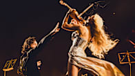 A man in black stands with outstretched arm as a woman with long blonde hair plays a violin above him, seen from behind.