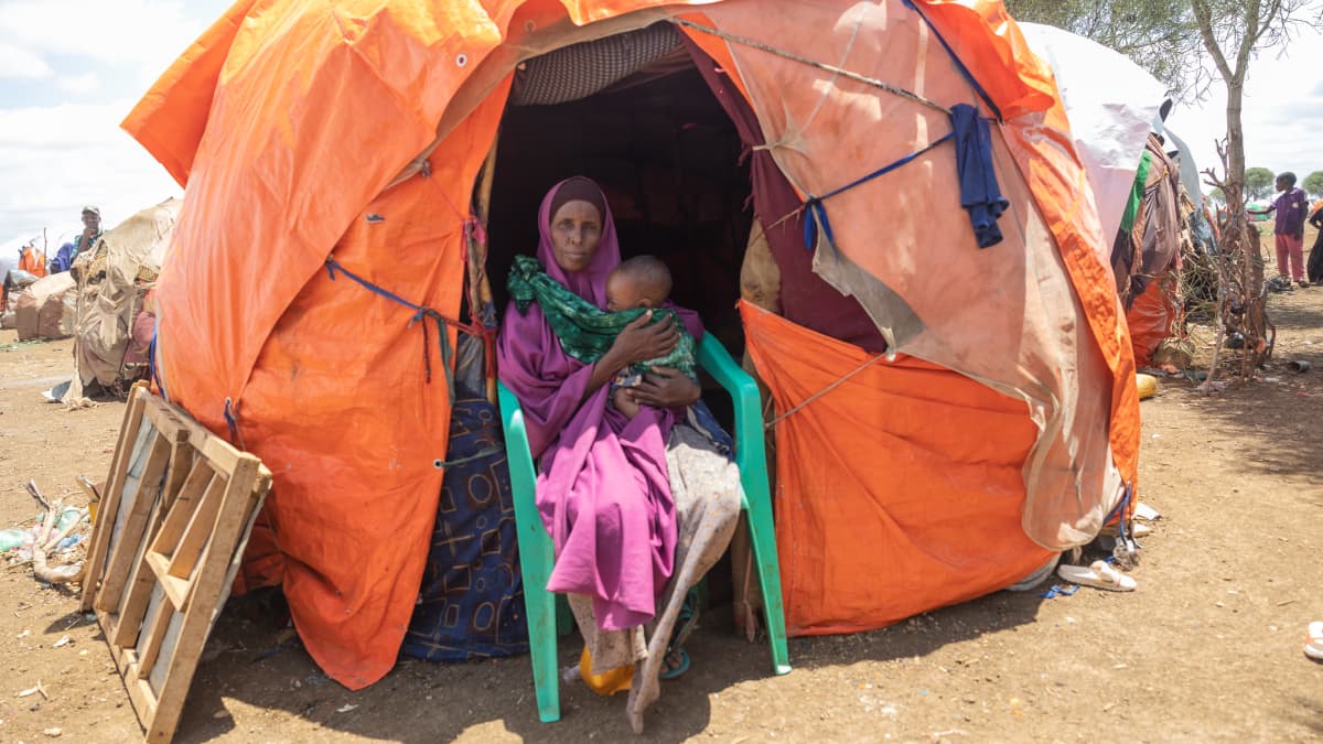 Somalialainen pakolaisäiti istuu oranssisilla pressuilla peitetyn majan suuaukossa muovituolillapakolaisleirillä, sylissään pieni vauva. 
