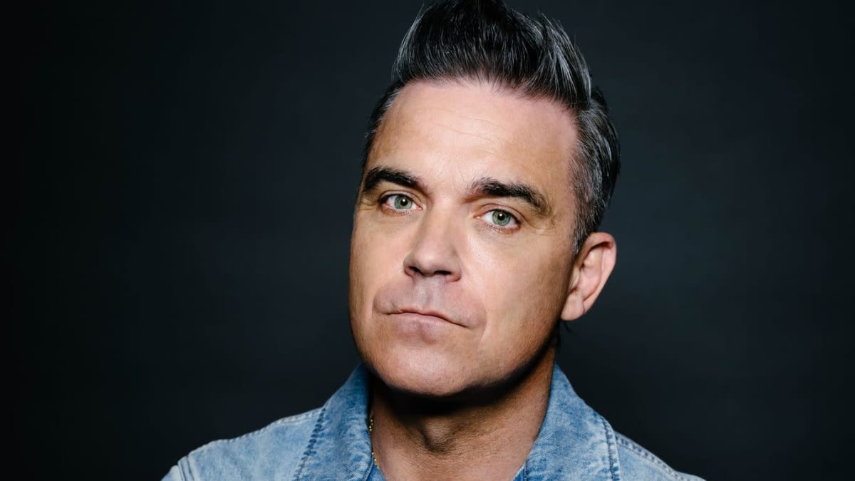 Robbie Williams seisoo tummaa taustaa vasten kädet puuskassa ja katsoo vakavana kameraan. Williamsilla on yllään farkkutakki.