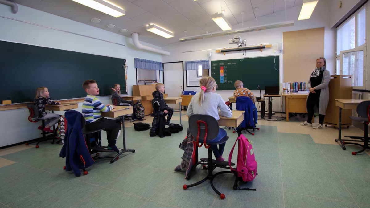 Pieni ryhmä kakkosluokkalaisia istuu pulpeteissaan, jotka on järjestetty turvavälietäisyyksille. Luokan edessä opettaja. 