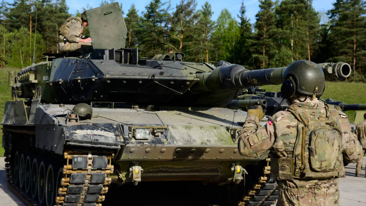 Kuvassa on Tanskan armeijan Leopard 2 -taistelupanssarivaunu. Vaunun edessä selin kameraan seisoo sotilas ja vaunun päällä on toinen sotilas. 