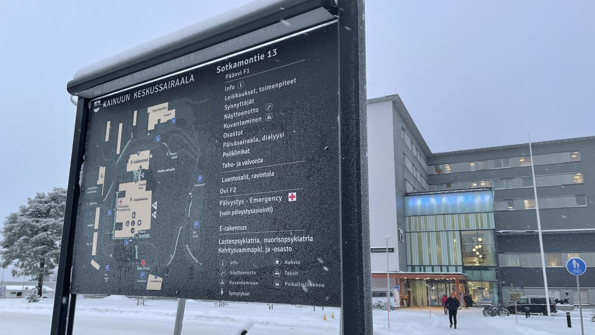Kyltti jossa kartta Kainuun keskussairaalasta, jossa lueteltu sairaalan osastot. Takana näkyy sairaalan sisäänkäynti.