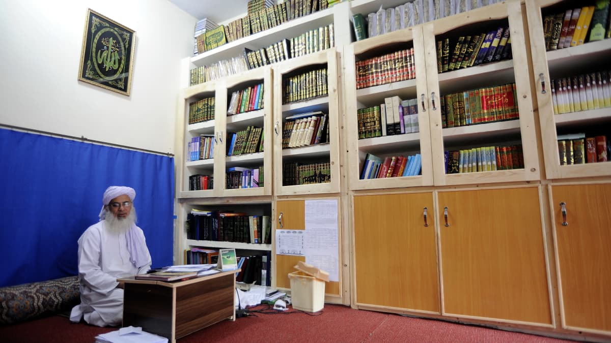 Parrakas silmälasipäinen mies istuu valkoinen turbaani päässään valkoisessa asussa kirjastomaisen huoneen lattialla pienen pöydän ääressä.