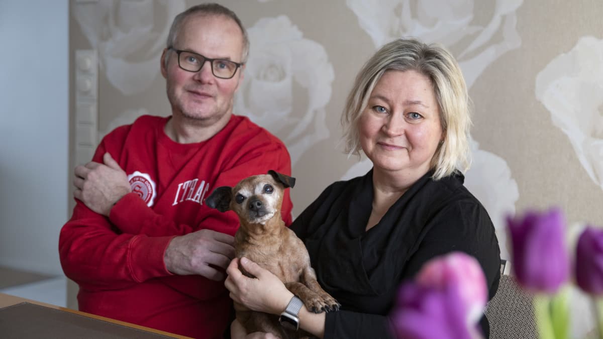 Jouko Haapalainen punaisessa paidassa istuu pöydän äärellä, vieressä vaimo Minna Haapalainen mustassa puserossa sylissään pieni kääpiöpinseri. 