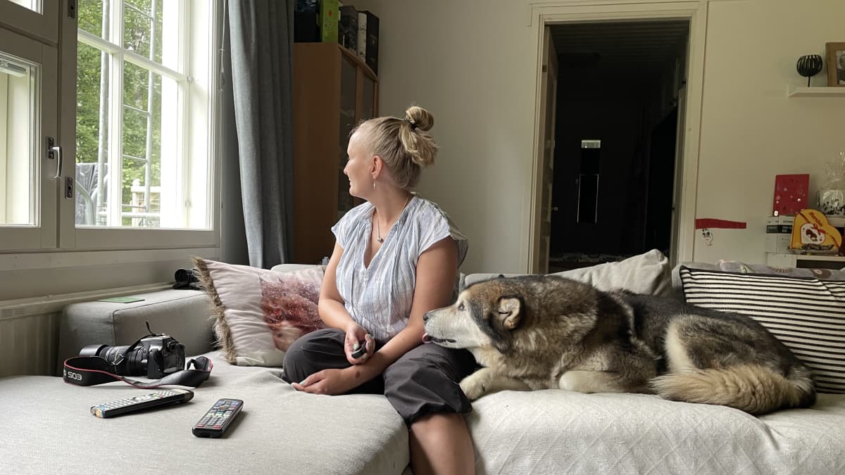 Ikkunasta ulos katsova nainen istuu sohvalla koiran kanssa kameran kaukolaukaisin kädessä. 