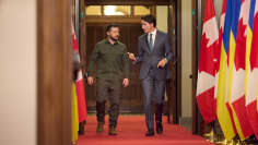 Zelenskyi ja Trudeau kävelevät vierekkäin käytävällä, jota reunustavat lippurivit.