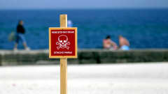 Varoituskyltissä punaisella pohjalla pääkallo, jossa varoitetaan Odessan rannan käyttäjiä miinoista. Taustalla meri ja rannalla istuvia ihmisiä.