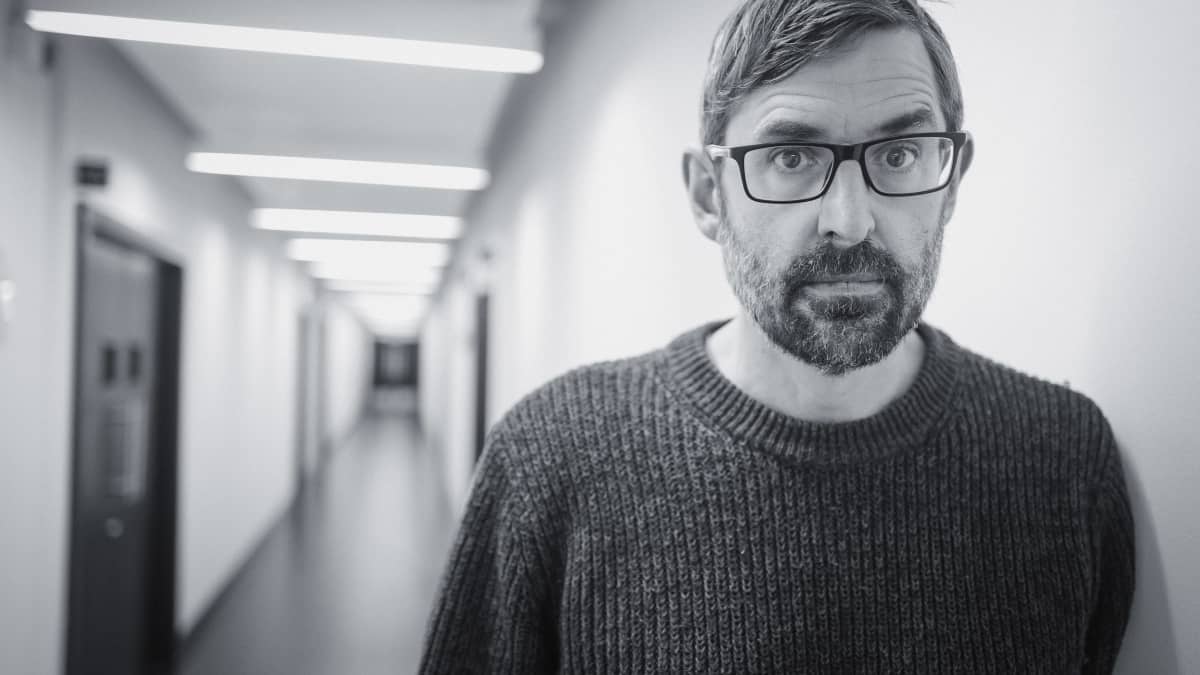 Louis Theroux mustavalkoisessa lähikuvassa pitkän käytävän etualalla.