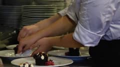 Suurkeittiön työntekijöiden kädet viimeistelevät jälkiruokalautasia, joilla suklainen jälkiruoka.