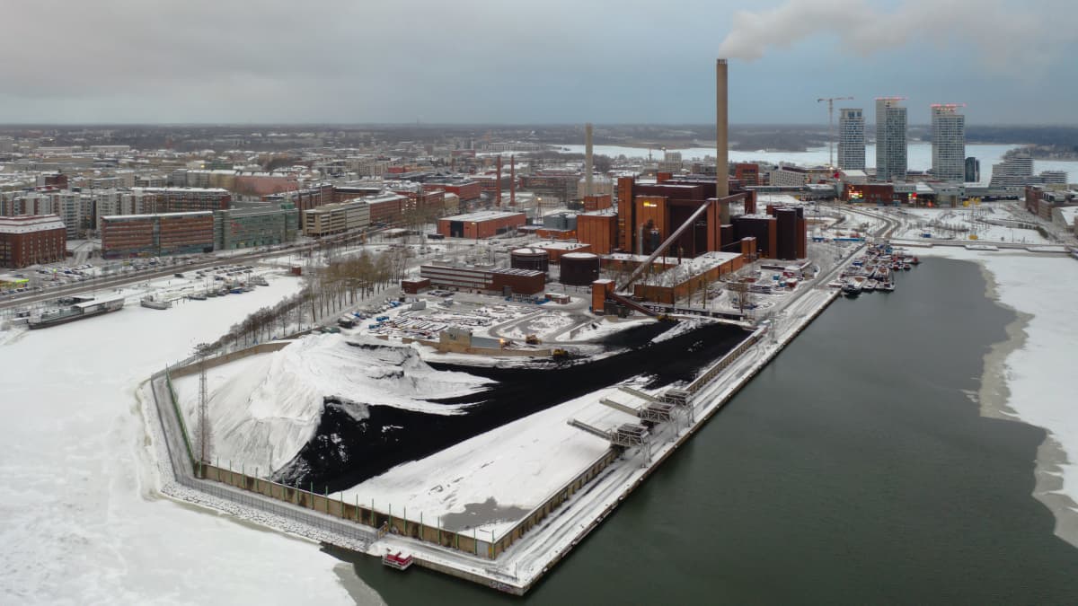 Ilmakuvassa Hanasaaren voimalaitos Helsingin Sörnäisissä. On talvi ja maa on valkoinen. Musta hiilikukkula on peittynyt osittain lumeen. Voimalaitoksen piipusta nousee harmaata savua. 