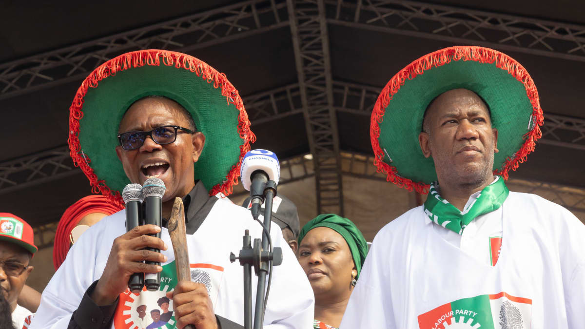 Two Nigerian politicians dressed in white robes and green top hats speaking in a stadium.