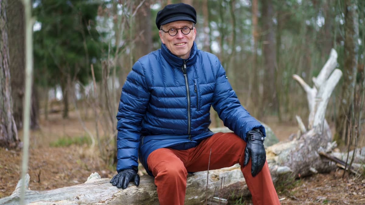 Maanmittauslaitoksen johtava asiantuntija Esa Ärölä istuu metsässä puunrungolla katsoen kameraan.