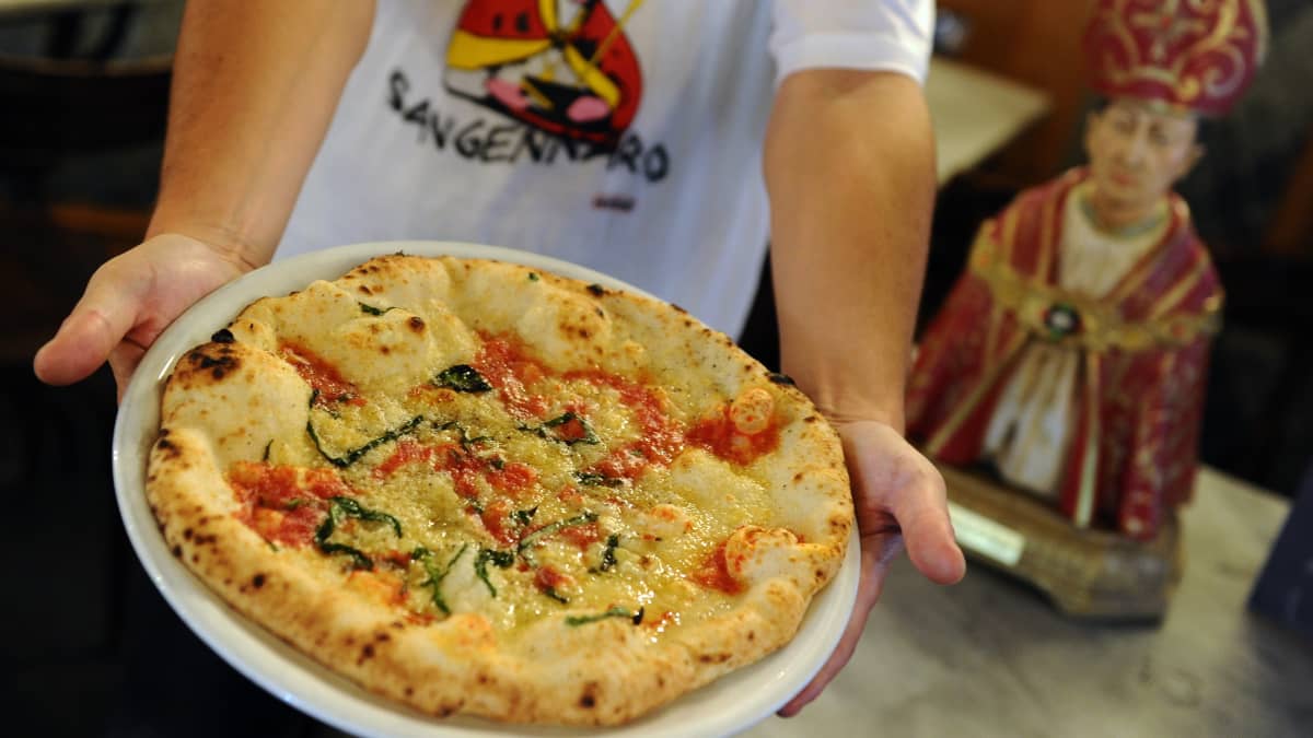 Mies ojentaa lautasta, jolla pizza, vieressään Napolin suojeluspyhimystä San Gennaroa esittävä pysti.