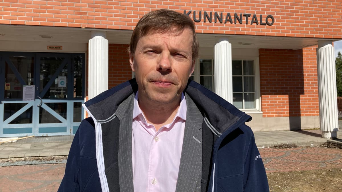 Timo Kurula katsoo kameraan silmiään siristellen ja takki auki, taustallaan Kittilän kunnantalo.