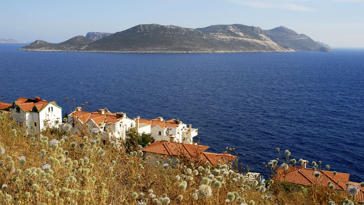 Maisema Turkin rannikolta kohti Kastellorizon saarta. Etualalla on taloja. Saari on keskellä sinisenä loistavaa merta.