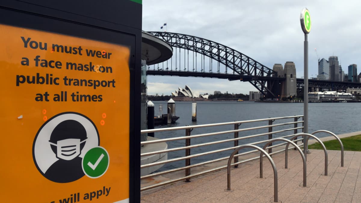 Kyltissä lukee: Julkisessa liikenteessä on aina käytettävä kasvomaskia sakon uhalla, taustalla Sydneyn keskustaa