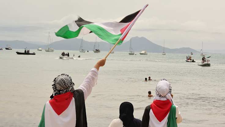 Three people seen from behind, wearing black, white, red and green robes, one waving a flag with the same colours at a dozen boats.