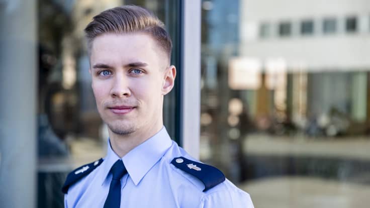 Nuori mies poliisin uniformussa katsoo kameraan. Taustalla lasinen ulkoseinä.