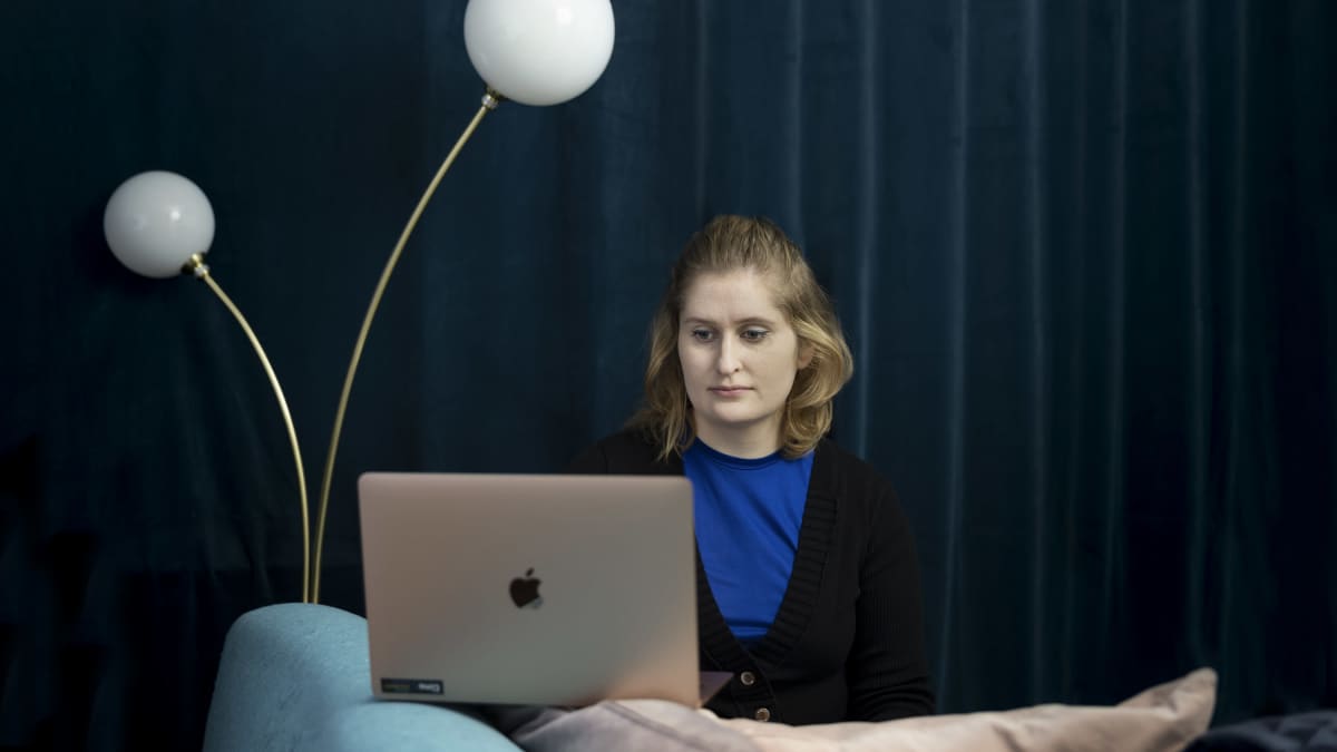 Liisa Salonen tekee töitä tietokoneella. Ikkunat on peitetty paksuilla verhoilla ja lattialla seisoo lamppu, josta kurkottaa kaksi pallokupua. 