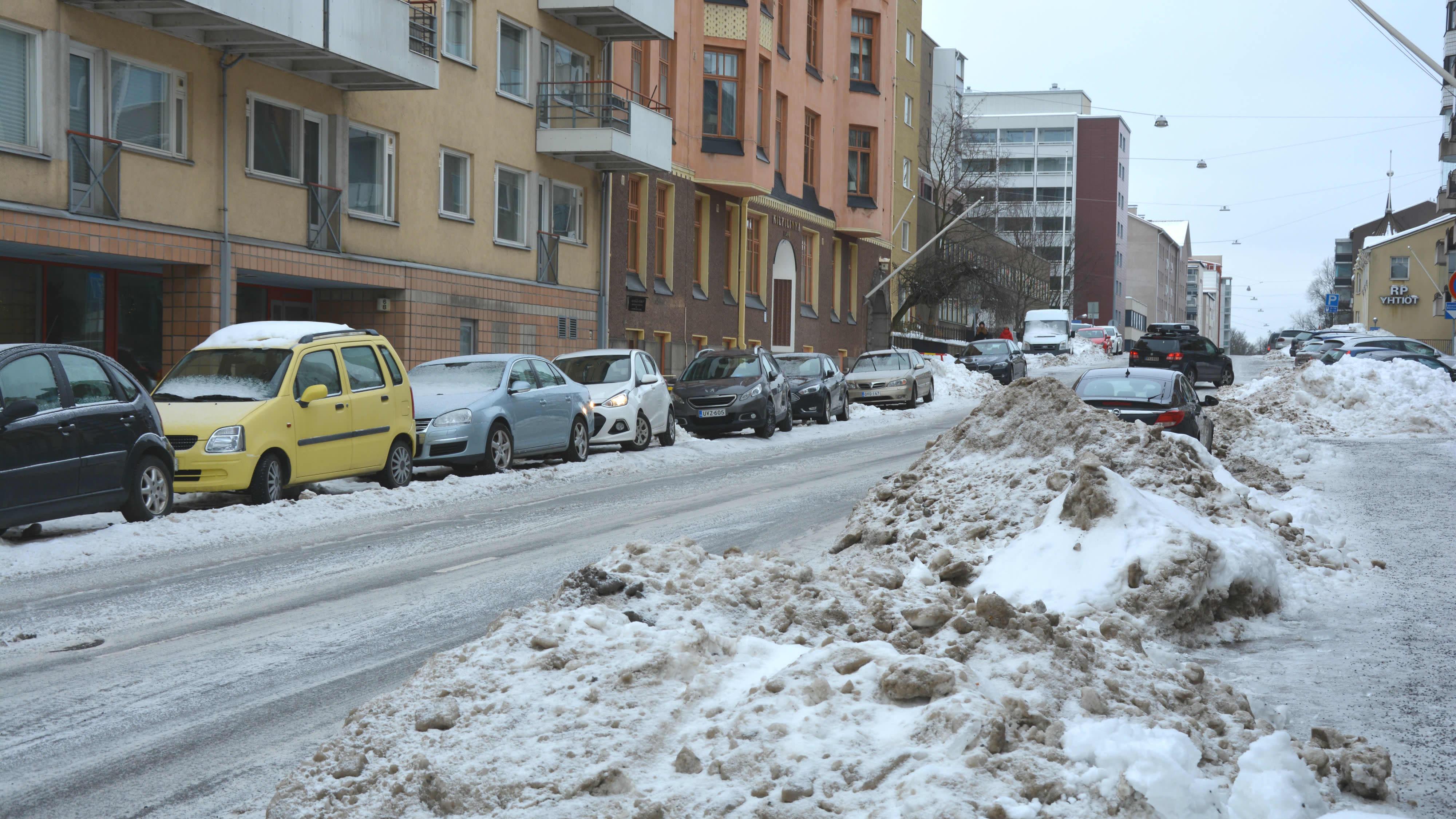 Stora snöhögar på en smal väg, flera bilar står parkerade längs vägen. 