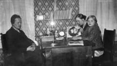  Lampaluodon ensimmäinen radio (kidekone). Radiota kuunnellaan saaristossa pöydän ääressä. Mies, tyttö ja lehteä lukeva nainen.