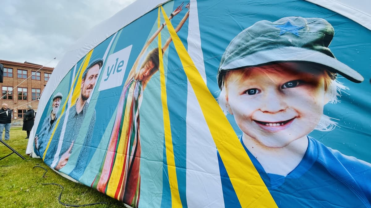 Turkoosi telttakangas, jossa hymyilevä pikkupoika ja muita ihmisiä sekä Ylen logo.