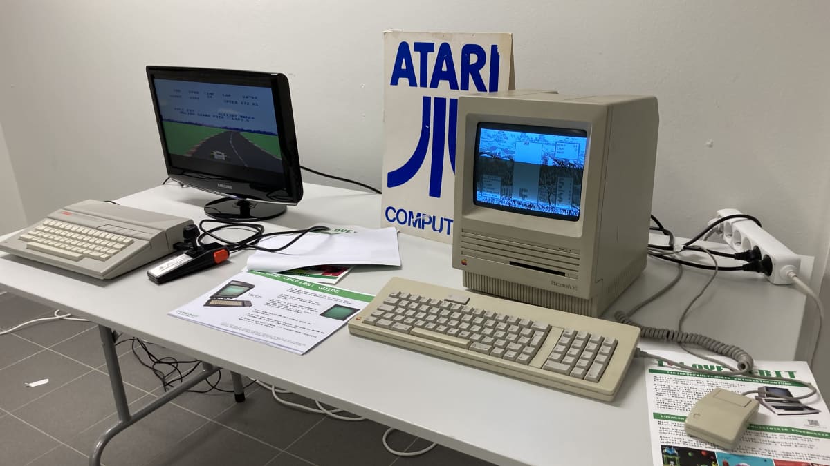 Kaksi vanhaa tietokonetta pöydällä. Taustalla pöydällä on kyltti, jossa on Atarin logo.
