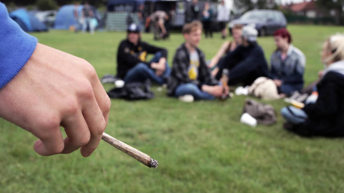 En hand håller i en joint i förgrunden. I bakgrunden syns suddigt ett gäng ungdomar som sitter i en ring på en gräsmatta.
