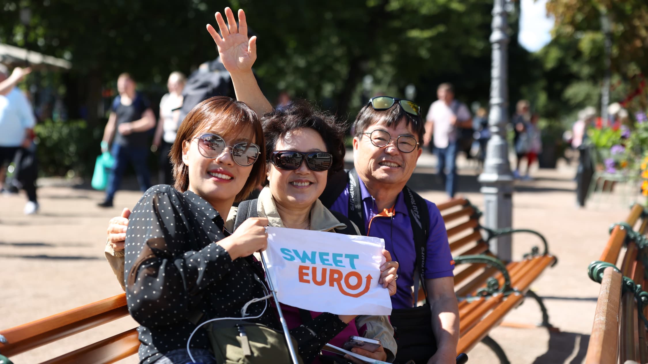 Etelä-Koreasta saapuneet kolme turistia istumassa Helsingin Espan lavan yleisöpenkillä ja katsomassa kameraan. Keskellä heillä on valkoinen lippu, jossa on tekstiä.