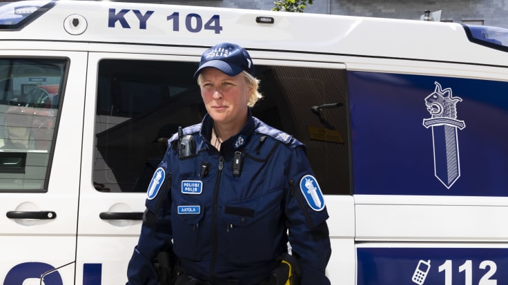 Sanna Aatola seisoo poliisin uniformussa poliisiauton edessä ja katsoo ohi kamerasta vakavan näköisin kasvoin.