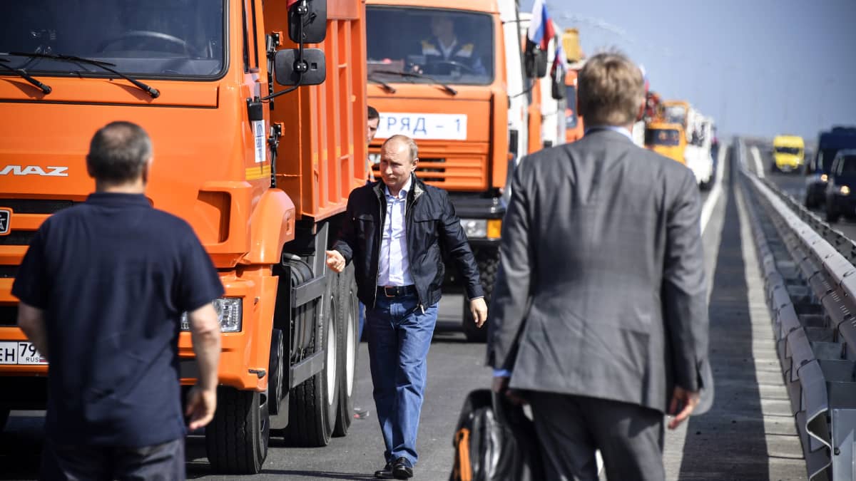 Vladimir Putin kävelee sillalla, taustalla jono kuorma-autoja.