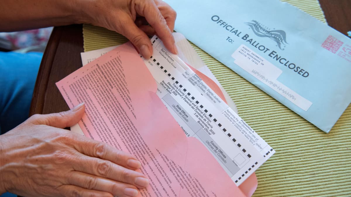 Lähikuvassa nainen sulkee vaalilippua postikuoreen. Kuoressa lukee englanniksi: Official Ballot Enclosed.