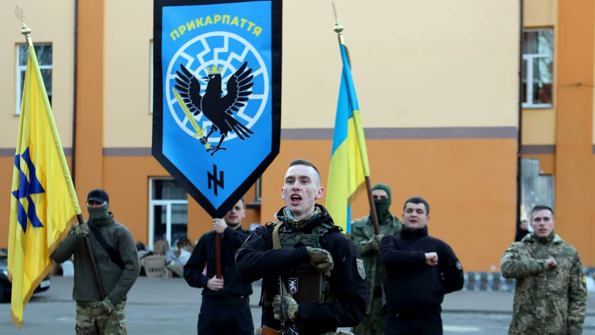 Sotilaspukuiset miehet seisovat rivissä oikean käsi nyrkissä rinnalla. Yksi kantaa Ukrainan lippua ja kahdella muulla on Azovin liikkeeseen viittaavat liput.