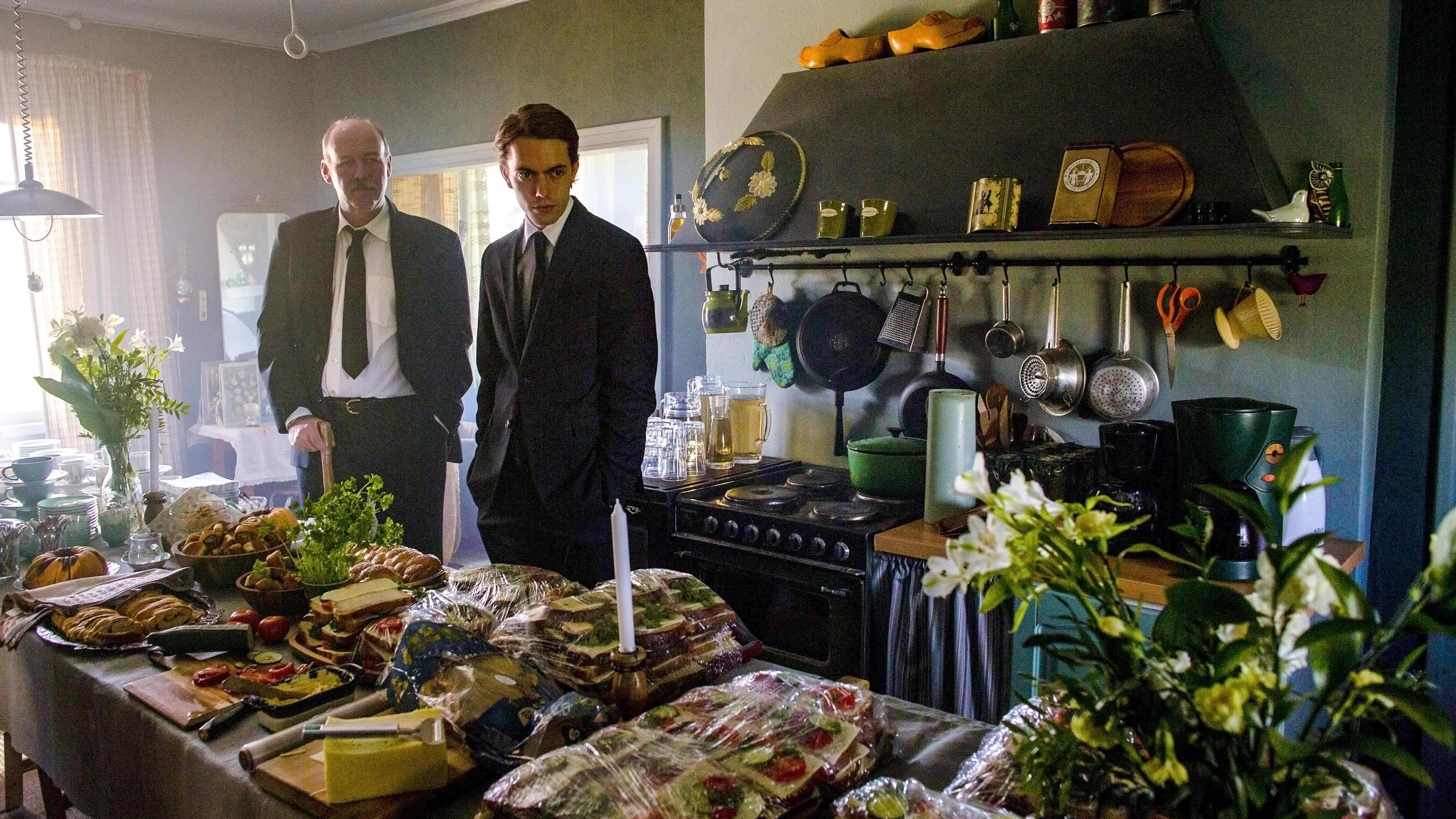 Nisse (Peik Stenberg) och Stefan (Martin Paul) står vid ett långbord fullt med smörgåsar och blommor.