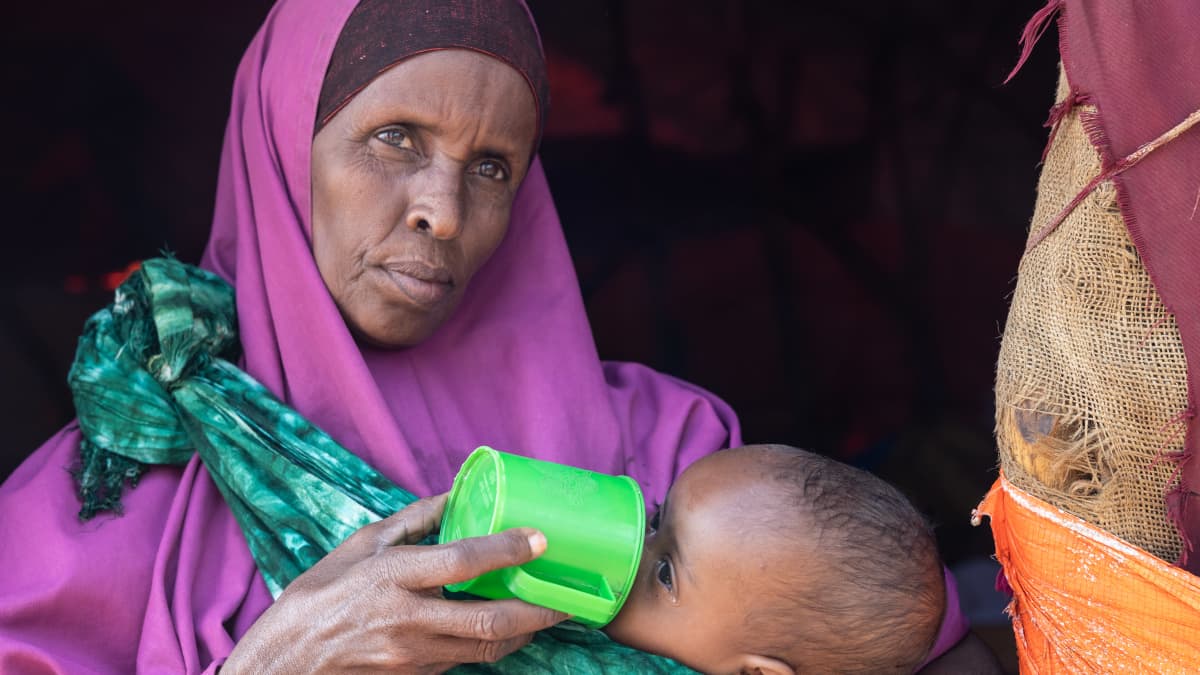 Somalialainen violettiin huntuun pukeutunut pakolaisäiti juottaa teltan oviaukossa pienelle vauvalle maitoa vihreästä muovimukista.