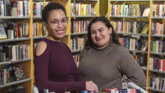 Aracelis Correa kuvassa vasemmalla ja Téri Zambrano oikealla poseeraavat kameralle kirjastosalissa. Aracelis nojaa käsivarttaan kuljetusvaunuissa oleviin kirjoihin. Taustalla näkyy kirjahyllyjä.