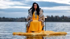 Kaija Koo istuu tuolilla veden keskellä Tuusulanjärven rannassa. Kaija Koolla on yllään keltainen mekko ja vaalea turkki.