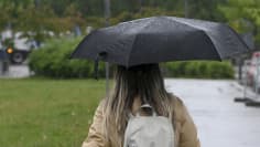 Takaapäin kuvattu pitkätukkainen henkilö kävelee sateenvarjon kanssa sadesäällä, taustalla näkyy nurmikkoa, asfalttia, puita ja pieni kaistale autotietä.