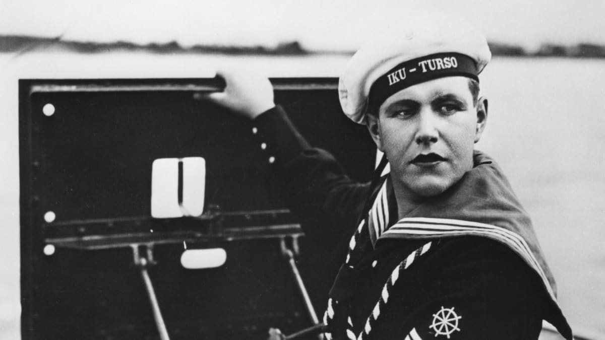 Georg Malmstén merimiespuvussa nousemassa kansiluukusta. Hatun nauhassa lukee Iku-Turso.