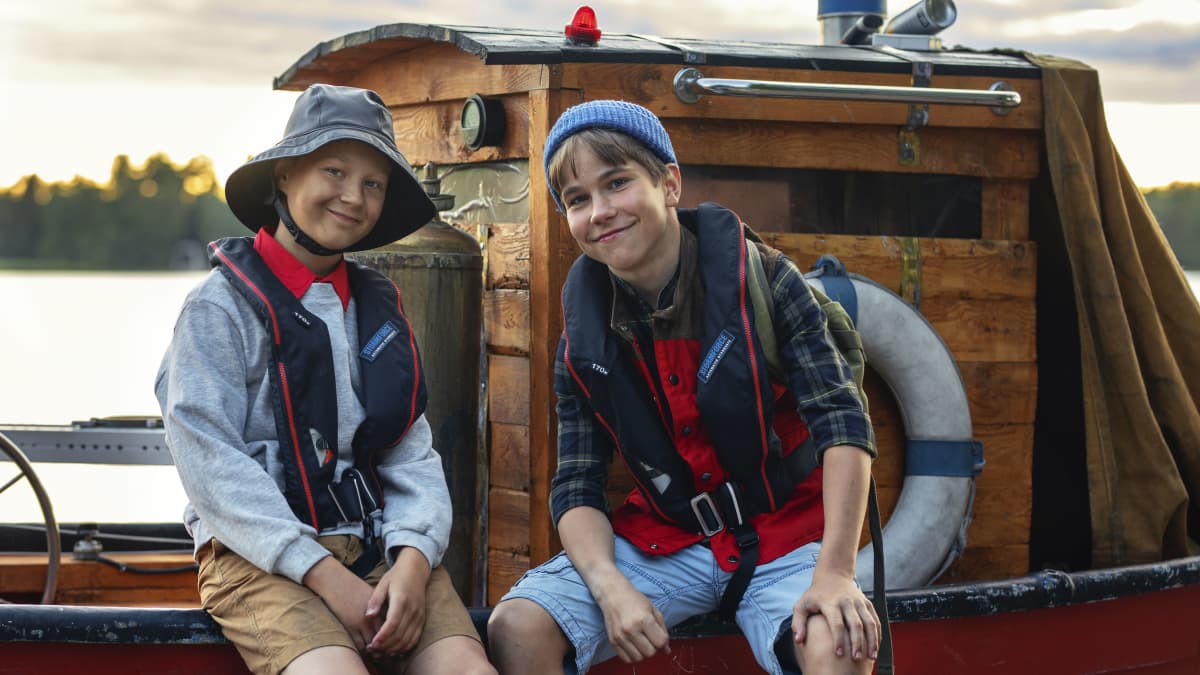 Kaksi poikaa istuu veneen laidalla, katsoo kameraan ja hymyilee.