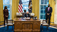 Joe Biden allekirjoittaa työpöytänsä äärellä. Kamala Harris ja Lloyd Austin katsovat vierestä.