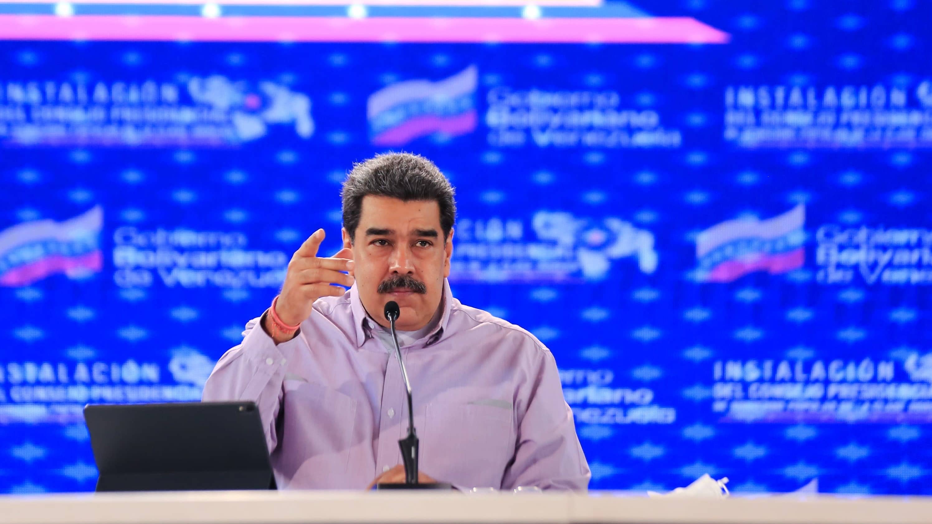 Maduro puhuu ja vahvistaa sanojaan kohottamalla oikeaa kättään. Hänellä on yllään vaalea kauluspaita. Hänen edessään pöydällä on mikrofoni ja näyttö. Taustakangas on sininen.