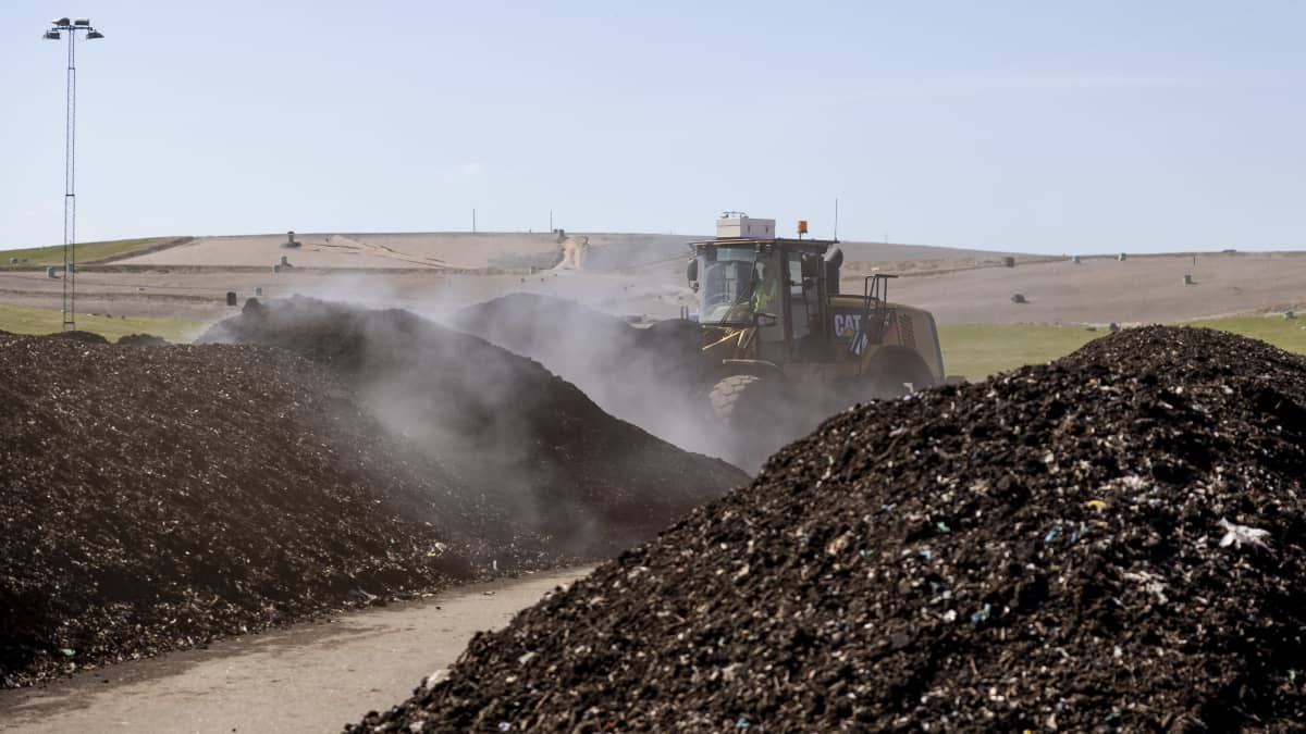Kuormaaja siirtelee hyöryäviä kompostikasoja. Taustalla maisemoitu kaatopaikkakukkula.
