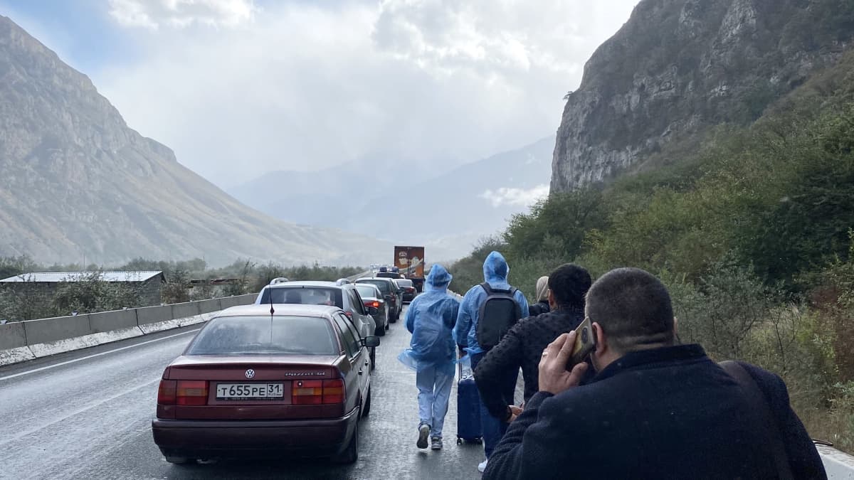 En grupp människor går bredvid en bilkö. Det regnar. I bakgrunden syns höga berg.
