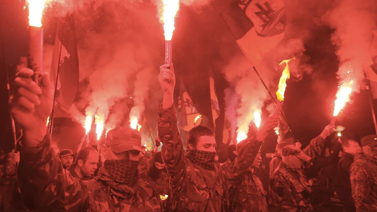 Maastopukuiset miehet pitävät soihtuja kädessään mielenosoituksessa. Taustalla näkyy Azovin rykmentin lippu.