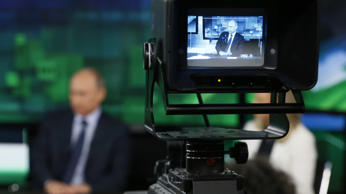 Presidentti Vladimir Putin näkyy häilyvänä kameran taustalla, RT-kanavan kuvaukset käynnissä.